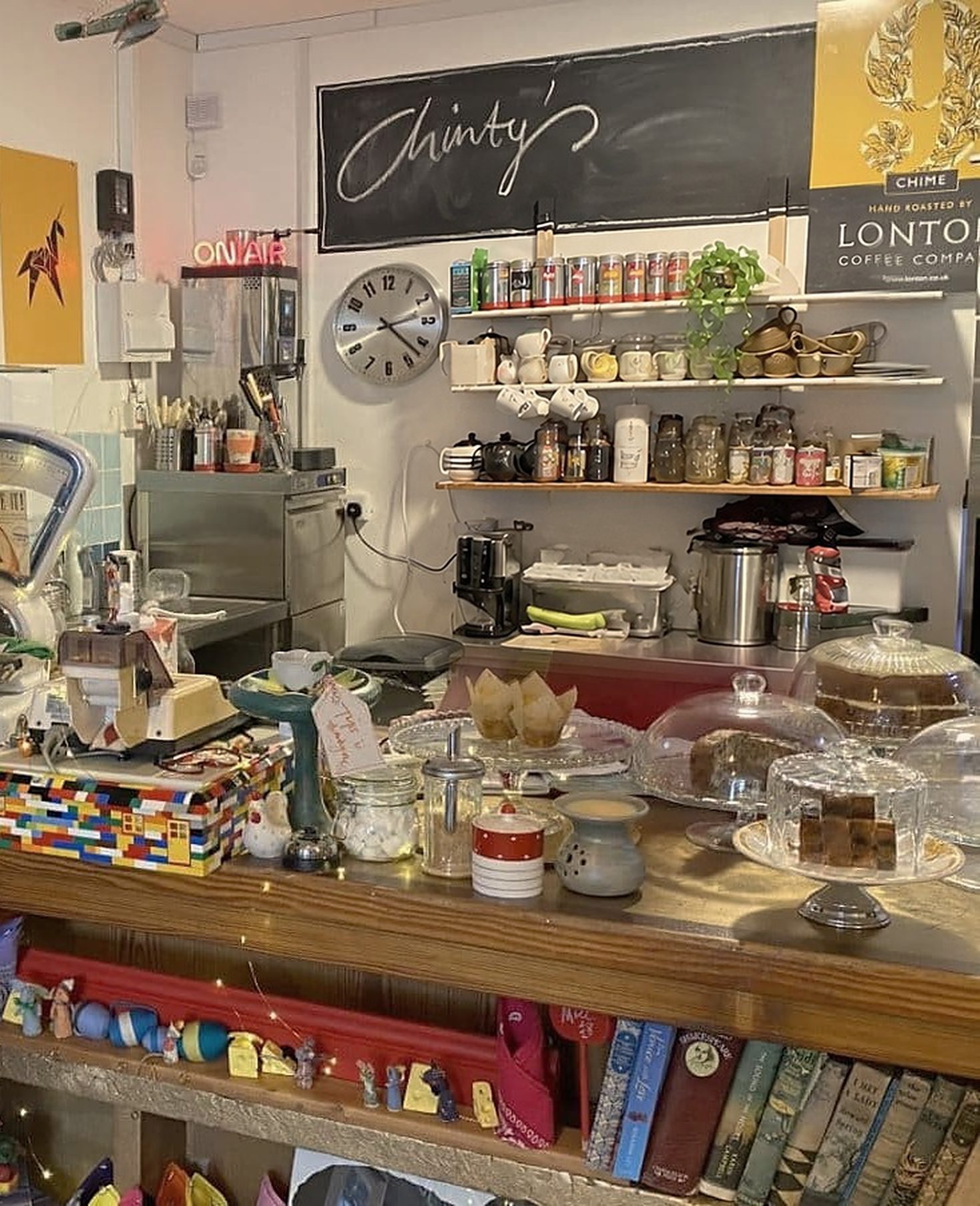 The counter: cakes under glass domes, a box built from toy bricks, books on the shelf below, and a chalkboard reading 'Chinty's' above shelves of tea and coffee