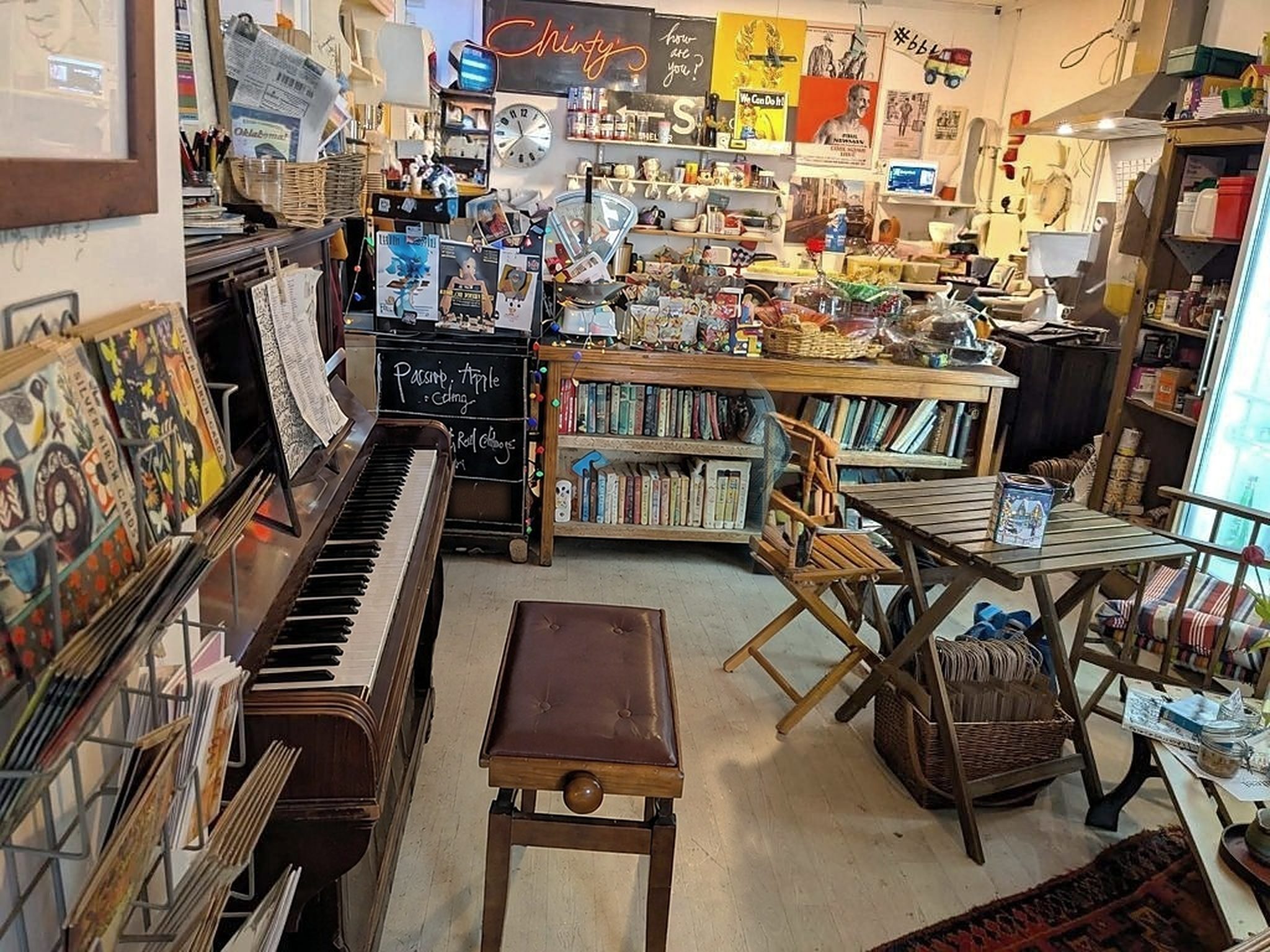 Inside Chinty's: an upright piano with an empty stool, wooden folding chairs and tables, bookshelves, a chalked soup board and shelves of jars and goods under a neon 'Chinty's' sign
