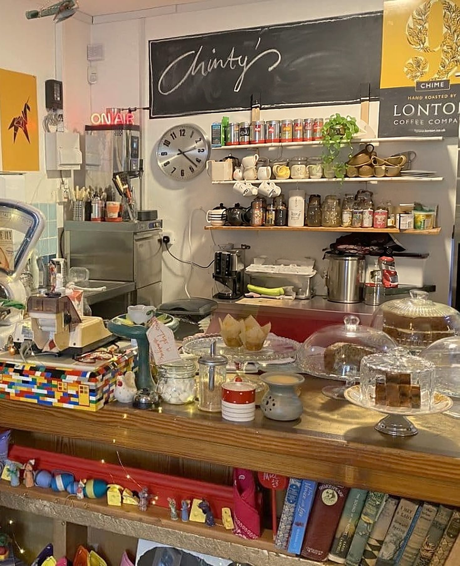 The counter: cakes under glass domes, a box built from toy bricks, books on the shelf below, and a chalkboard reading 'Chinty's' above shelves of tea and coffee