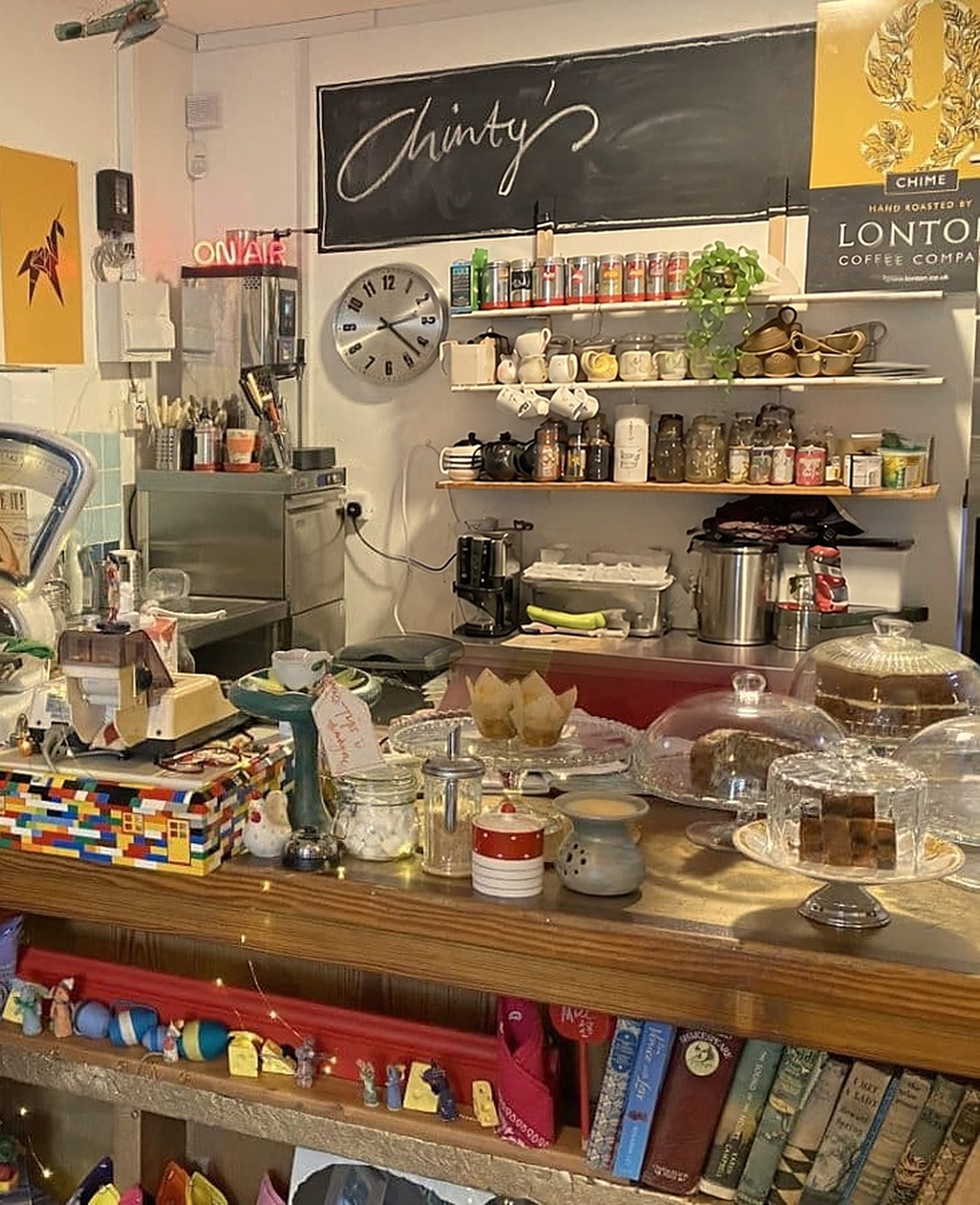 The counter: cakes under glass domes, a box built from toy bricks, books on the shelf below, and a chalkboard reading 'Chinty's' above shelves of tea and coffee