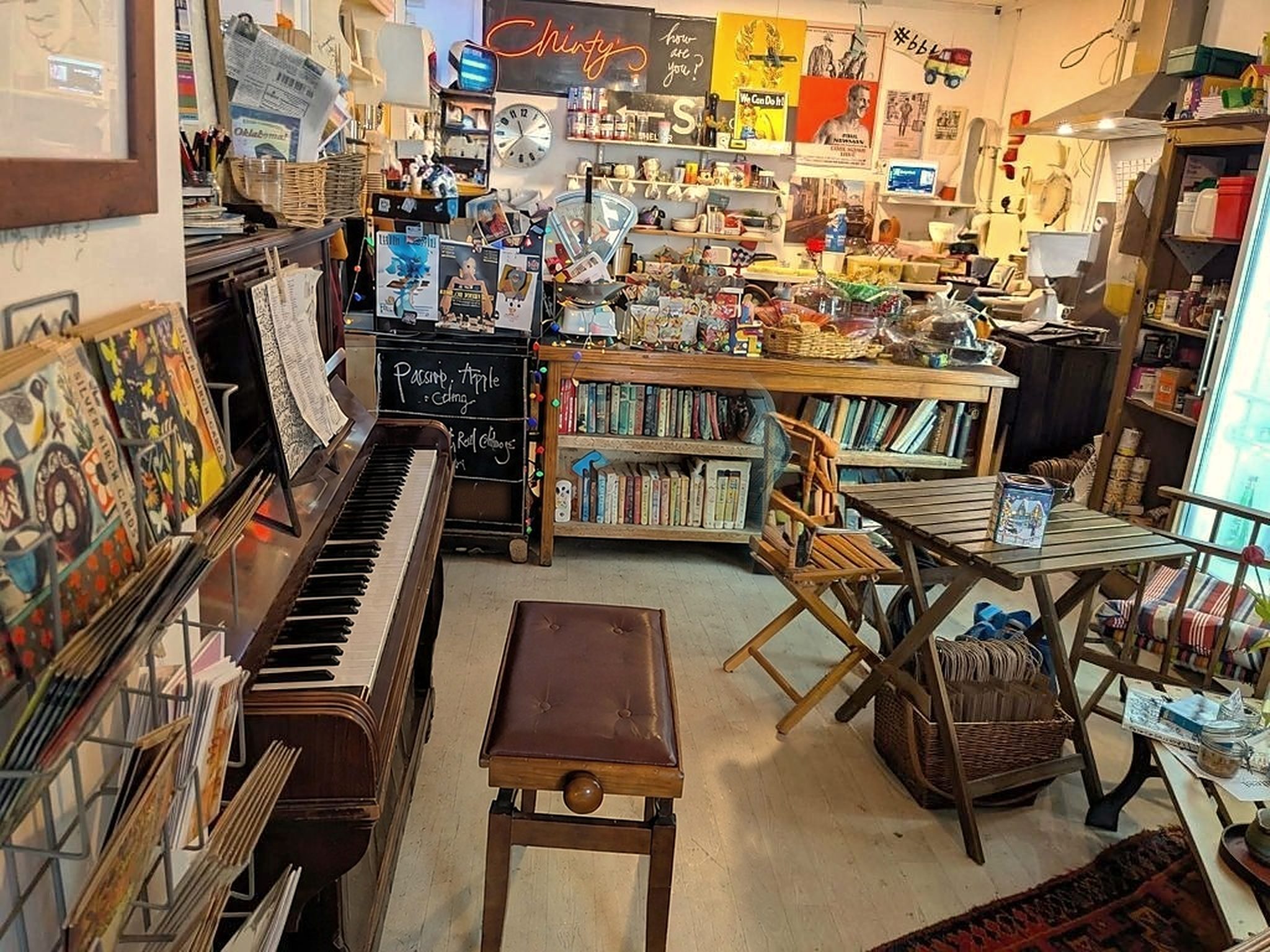 Inside Chinty's: an upright piano with an empty stool, wooden folding chairs and tables, bookshelves, a chalked soup board and shelves of jars and goods under a neon 'Chinty's' sign